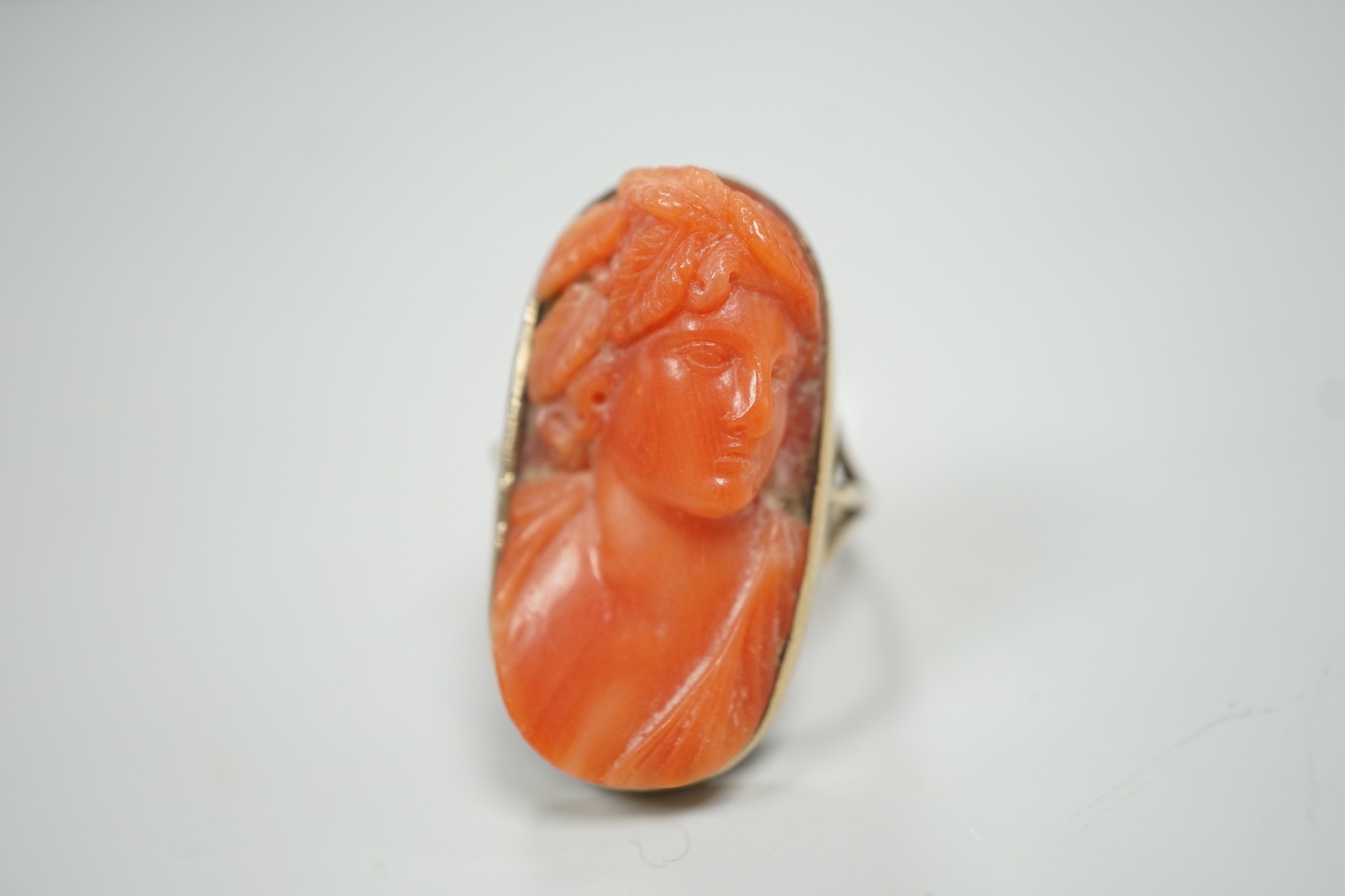 A yellow metal and coral set oval dress upfinger ring, the coral carved with a classical bust to sinister, size O, gross weight 8.2 grams.